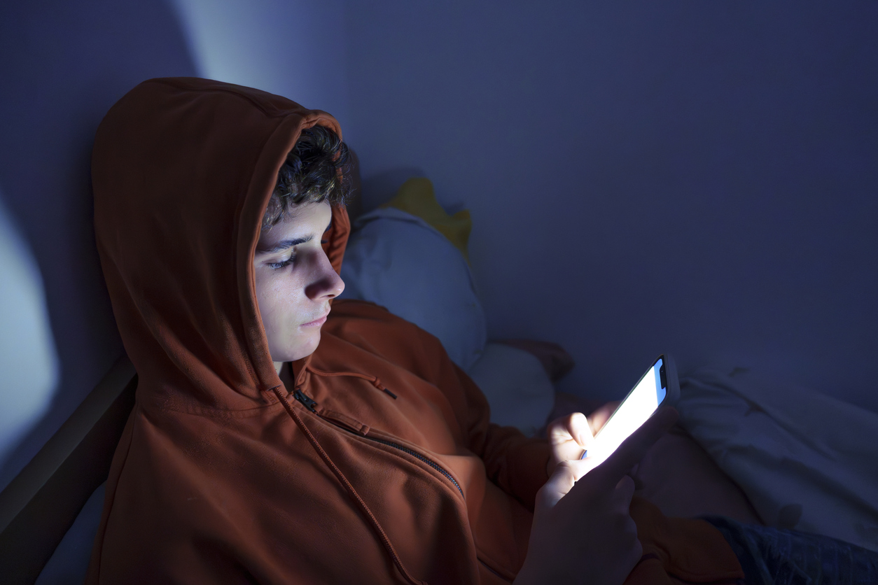 jongen ligt op bed in het donker en bekijkt sociale media op telefoon