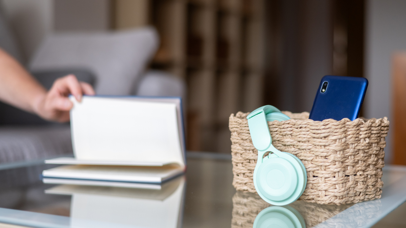 Smartphone en koptelefoon liggen in een mand terwijl iemand op de achtergrond een boek leest.